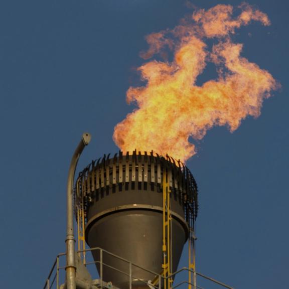 Industrial flare stack burning excess gas against a clear blue sky.