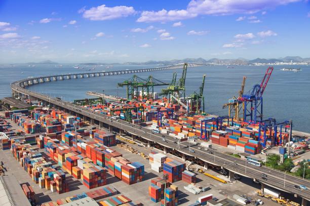 Vista ampla de porto de cargas com contêineres, guindastes e ponte sobre o mar
