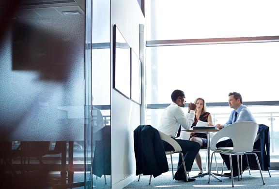 Three business professionals having a focused discussion around a table in a modern office meeting room with natural light.