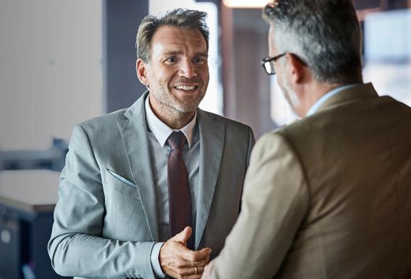 Two professional businessmen in conversation, smiling and shaking hands in a corporate office
