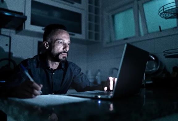 Homem trabalhando à noite no laptop em home office, fazendo anotações em papel sob pouca luz