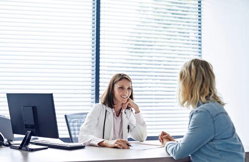 Female doctor sitting at desk smiling while consulting with woman patient in office with computer and blinds in background