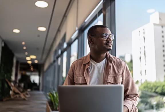 Homem com laptop sentado em escritório moderno, olhando pela janela pensativamente.