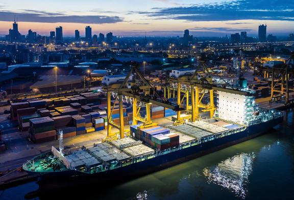 An illuminated cargo ship being loaded with shipping containers at a bustling port terminal at dusk, with cranes in operation and a city skyline in the background.