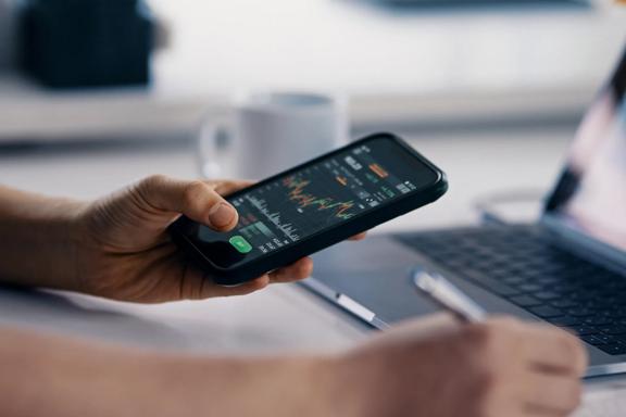 Hand holding smartphone showing trading platform with laptop in background