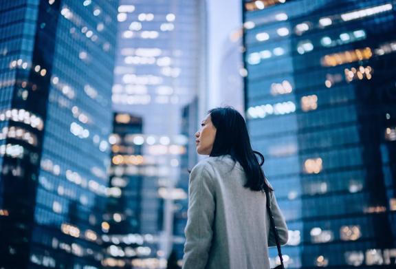 Businesswoman in a gray coat looking up at illuminated skyscrapers in a modern city at dusk.
