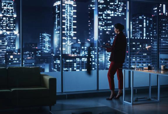 Businesswoman standing by window in high-rise office at night, using digital tablet with city lights in background.