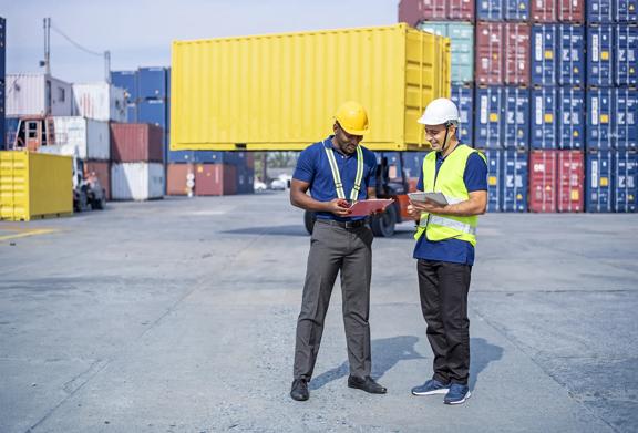 Dois trabalhadores com equipamentos de segurança discutindo logística perto de contêineres de carga em um pátio de embarque.