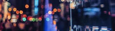 A blurred city street at night with colourful bokeh lights and an illuminated digital display screen in the foreground.