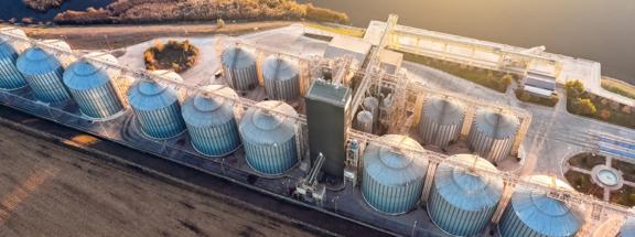 Vista aérea de uma instalação industrial com grandes silos e uma planta de processamento ao lado de um terreno aberto e água