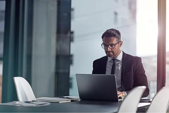 Executivo usando terno escuro e camisa branca, trabalhando em notebook sobre mesa de reunião com cadeiras brancas e parede de vidro ao fundo