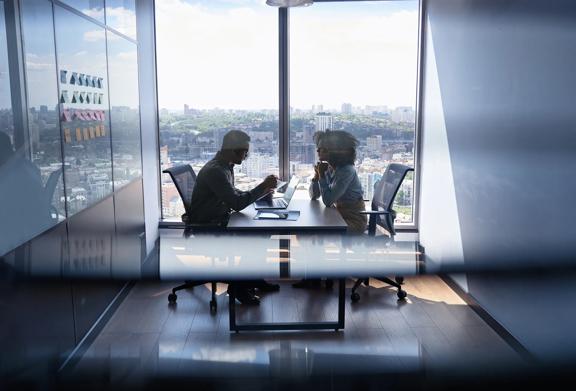 Two professionals having a focused discussion in a modern office meeting room with large windows and city skyline view.