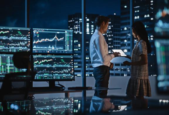 Two colleagues standing looking at a laptop by a window in a dark office