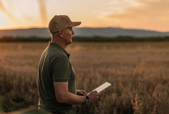 Farmer holding a tablet while observing a crop field at sunset, symbolizing precision agriculture and digital farming solutions.