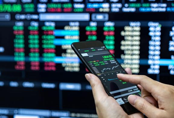 Close-up of hands using a smartphone app to monitor stock market data with trading screens in the background