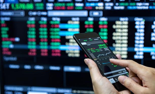 Pessoa usando um smartphone para acompanhar dados do mercado de ações ao vivo em frente a uma tela de negociação