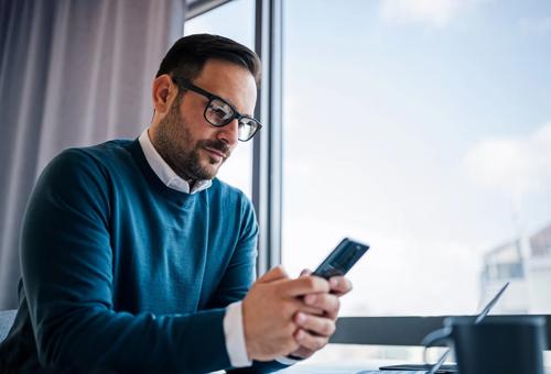 Professional man in glasses using smartphone at modern office desk with laptop and coffee