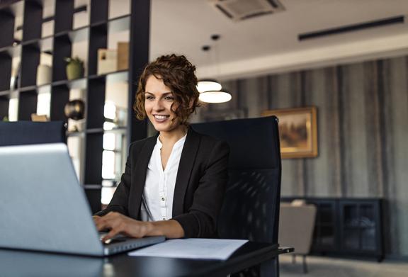 Confident businesswoman smiling while working on a laptop in a modern office environment.