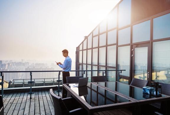 Businessman using smartphone on rooftop office terrace with panoramic city view, modern glass building and outdoor meeting table.