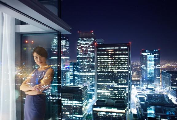 Confident businesswoman standing by high-rise window at night with illuminated city skyline in background.