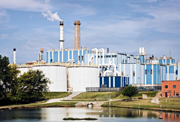 Grande instalação industrial azul e branca com chaminés de fumaça ao lado de um lago e árvores