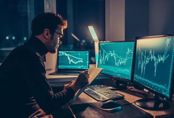 Financial analyst reviewing market trends and taking notes in front of multiple monitors at night