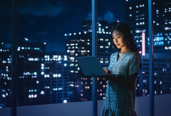 Woman holding laptop standing by window in dark office