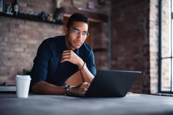 Homem jovem com óculos, apoiado na mesa de escritório moderno, usando laptop para trabalho corporativo, com café ao lado.