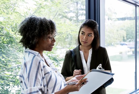 Two professional women having a thoughtful discussion near a window, with one reviewing notes on a clipboard.

