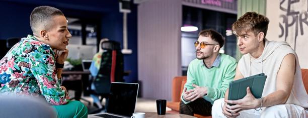 Three people seated in a collaborative office setting, engaged in discussion with one holding a tablet, surrounded by desks and computer monitors.