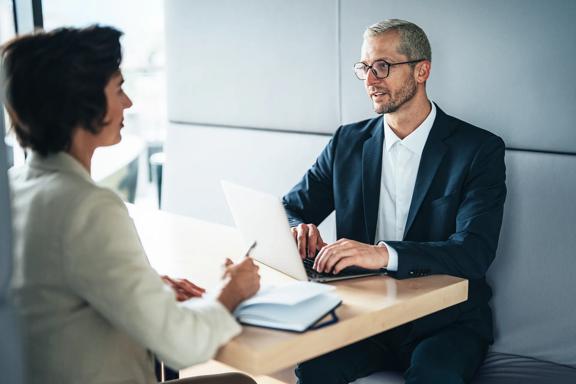 Dois executivos em reunião de negócios, sentados à mesa com laptop e caderno, discutindo estratégias e planejamento corporativo.