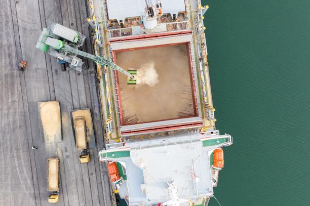 Visão aérea de navio cargueiro sendo carregado com grãos em terminal portuário com caminhões e guindaste