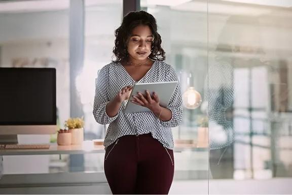 Pessoa segurando tablet branco em pé, vestindo camisa de estampa geométrica e calça vinho, em escritório com parede de vidro e mesa com computador ao fundo