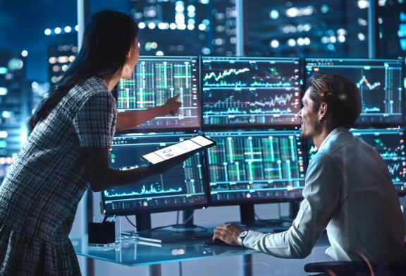 Two financial professionals analyzing stock market charts on multiple monitors in a modern office at night