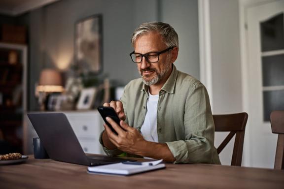 Person sitting at a table using a smartphone while working on a laptop in a home office setting.