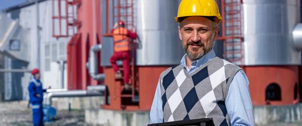 Trabalhador do sexo masculino usando um capacete amarelo e segurando um tablet, com equipamentos industriais e trabalhadores ao fundo