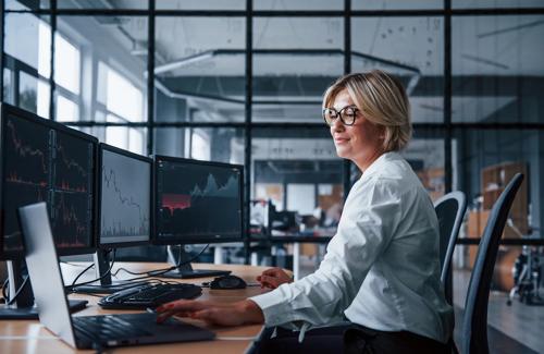Woman working at trading desk with multiple monitors displaying financial market charts and data