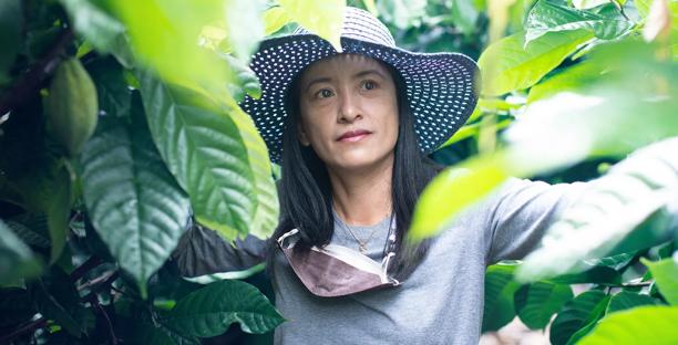 Woman wearing a wide-brimmed hat looking thoughtfully through foliage, symbolizing sustainable agriculture or environmental stewardship.