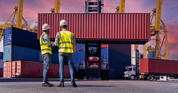 Two workers in safety vests and helmets overseeing container loading operations at a busy shipping port