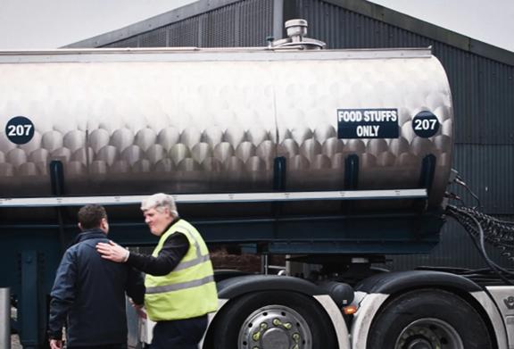 Food-grade stainless steel tanker truck labeled "Food Stuffs Only" at a logistics facility, with two workers onsite