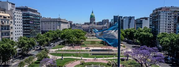A wide-angle view of the Argentine National Congress building in Buenos Aires, seen from Plaza del Congreso with an Argentine flag in the foreground and jacaranda trees in bloom.