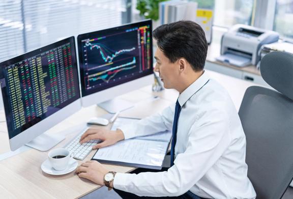 Person working at a desk with two large monitors displaying financial charts, stock prices, and market data. A laptop, documents, and a cup of coffee are also on the desk in a modern office setting.