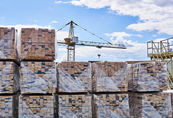 Stacked bundles of wood wrapped in plastic with a crane in the background at a lumber yard.