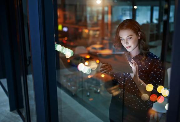 Woman working late at night, using a tablet with city lights in the background.