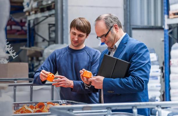 Engineer and manager inspecting orange mechanical parts in a warehouse or manufacturing facility.